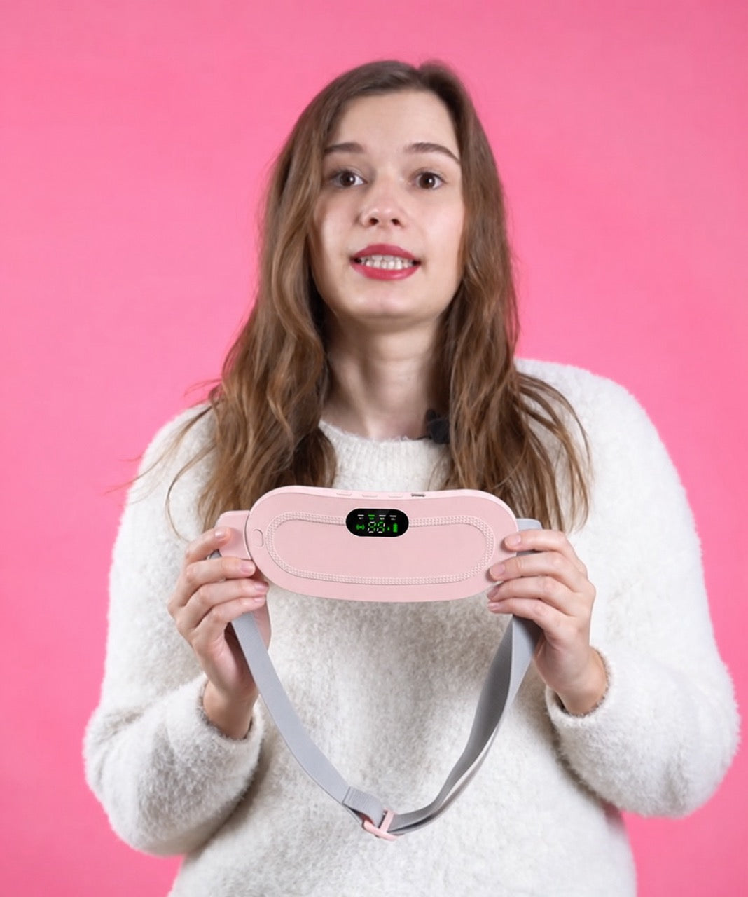 A woman with long brown hair wearing a white fuzzy sweater holds a pink digital massager with a gray strap against a solid pink background.