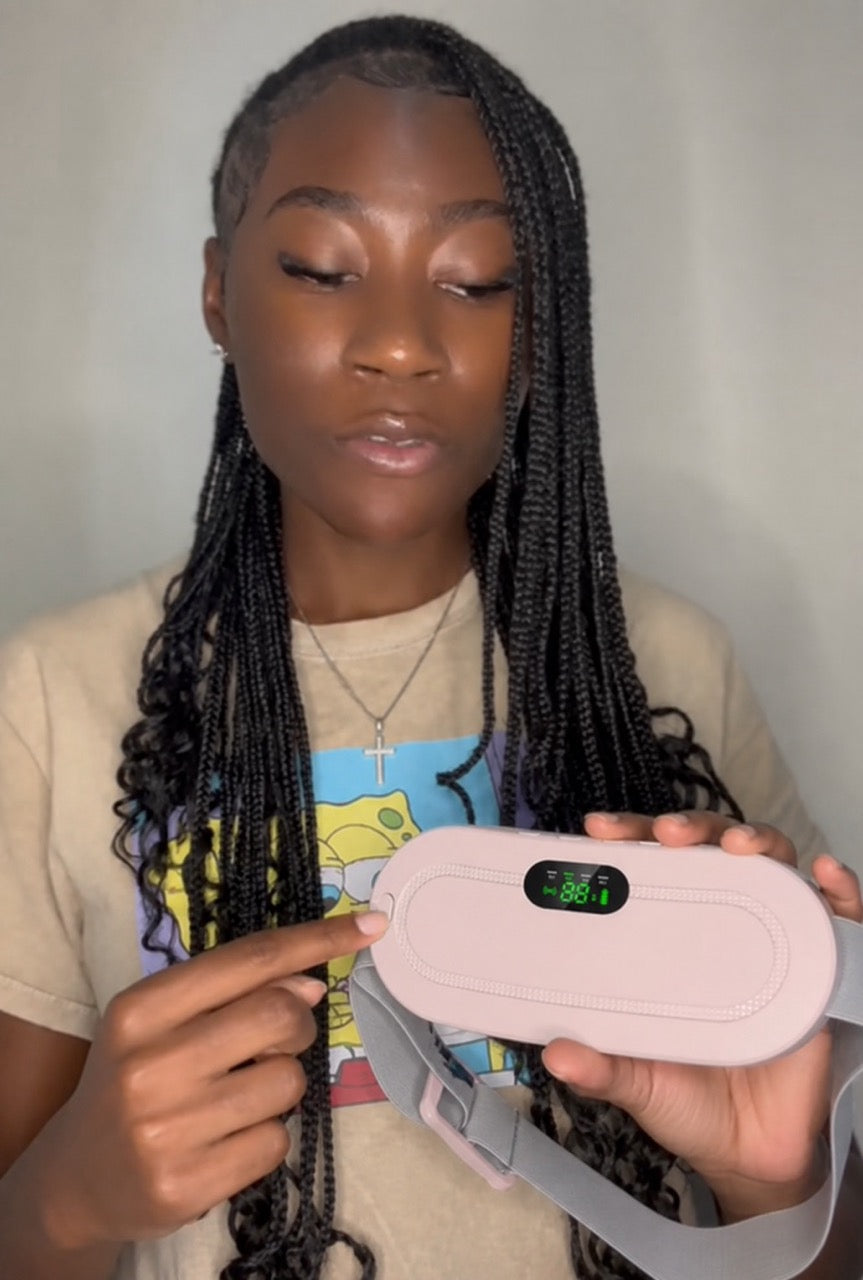 A woman with long black braids holds a light pink electronic device with a digital display screen, wearing a beige graphic t-shirt against a plain light gray wall.
