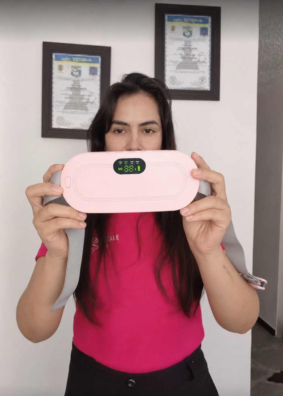 A woman wearing a bright pink t-shirt holds up a light pink electronic device with a digital display screen, set against a white wall with two framed certificates.